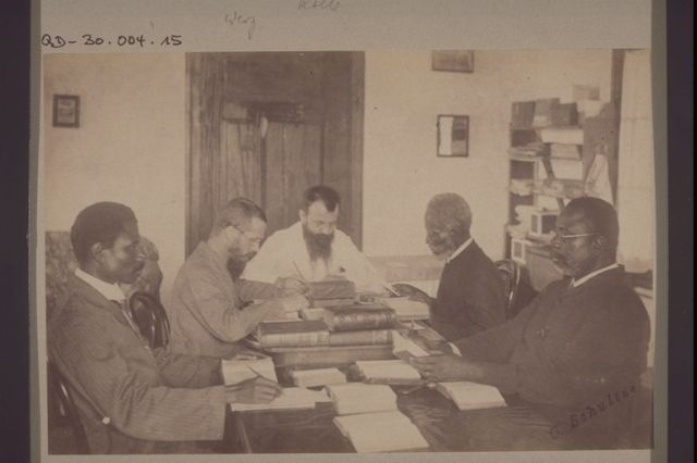 Historische Aufnahme von fünf Männern an einem Tisch, die aufgeschlagene Bücher vor sich haben. Die vorderen drei Figuren haben schwarze Haus, am Tischende sitzen Weisse.