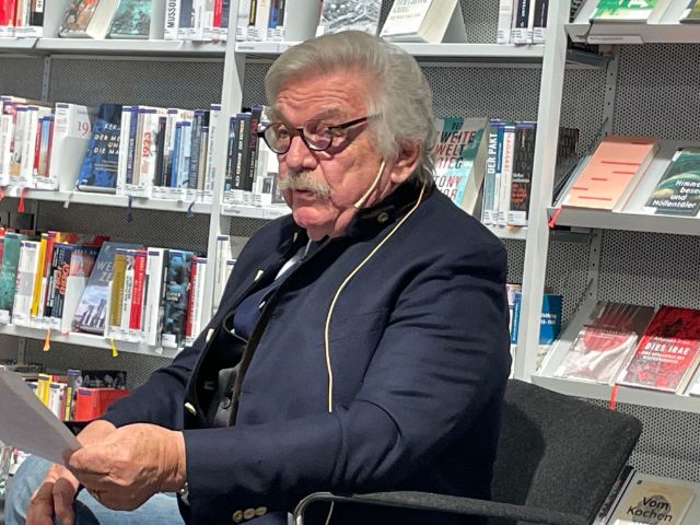 Ein älterer Herr mit Brille, grauem Schnauz und blauem Kittel liest etwas vor 
