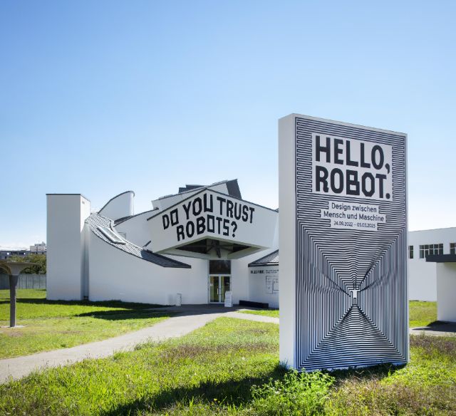 Abstrakt gebautes Gebäude; vor Gebäude Plakat von Ausstellung Hello Robot.