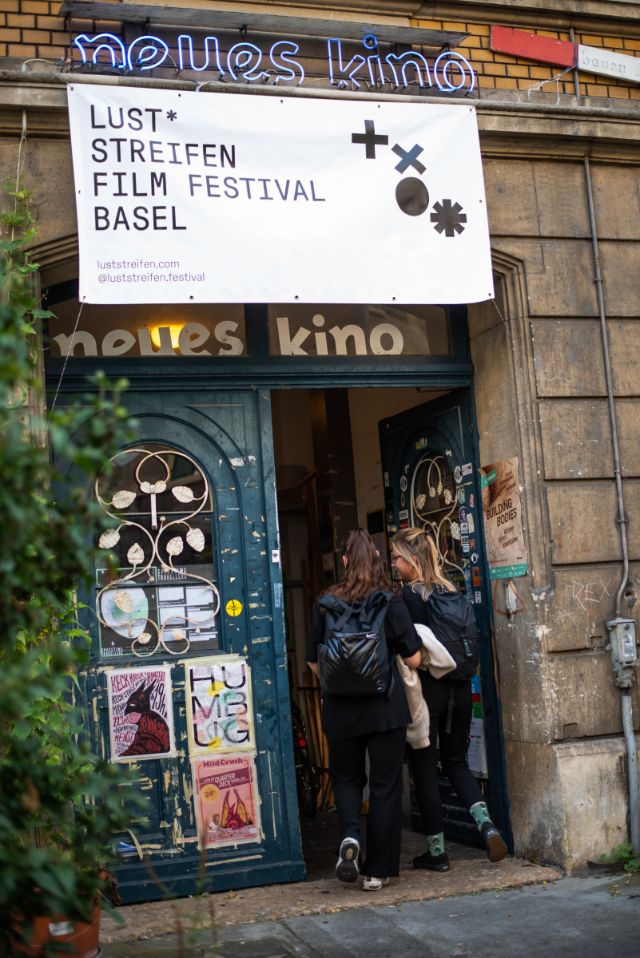 Eingang des neuen Kinos, zwei Personen gehen zur Tür rein; über der Tür hängt ein Plakat des Luststreifen Film Festivals