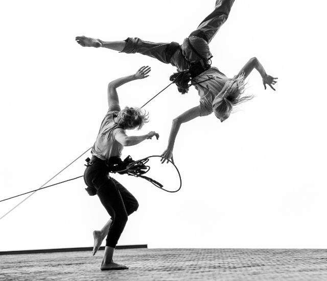 zwei Artist:innen mit langen Seilen tanzen auf einer senkrechten Backstein-Fassade 